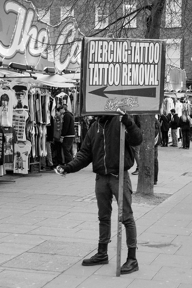 © Neil Turner, January 2014. Man holding sign offering tattoos an tattoo removal touts for business on the pavement just outside one of main areas of Camden Market