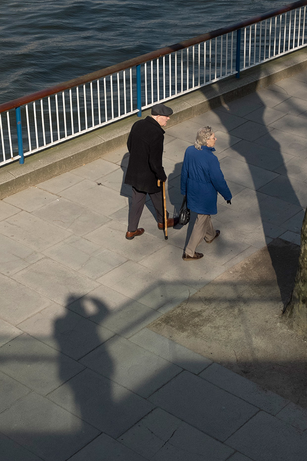 ©Neil Turner, March 2014. The photographer's shadow forms part of the scene as an elderly couple walk along South Bank of the River Thames in London.
